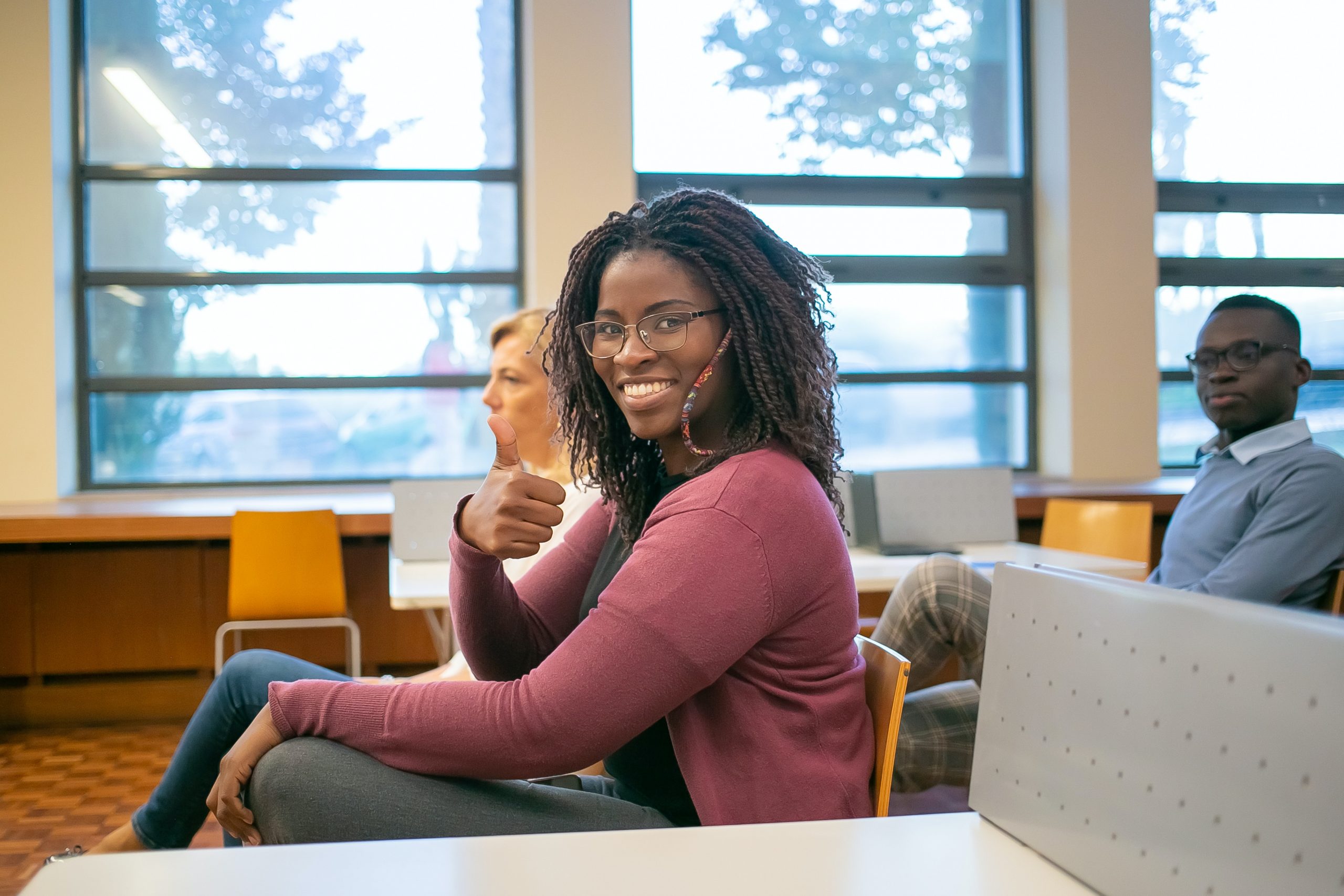 A black student in a red cardigan and glasses gives a thumbs-up to the camera